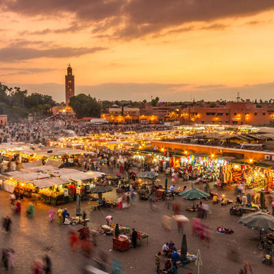 Drukke Djemaa el Fna-plein in Marrakech bij zonsondergang, met marktstalletjes en de Koutoubia-minaret.
