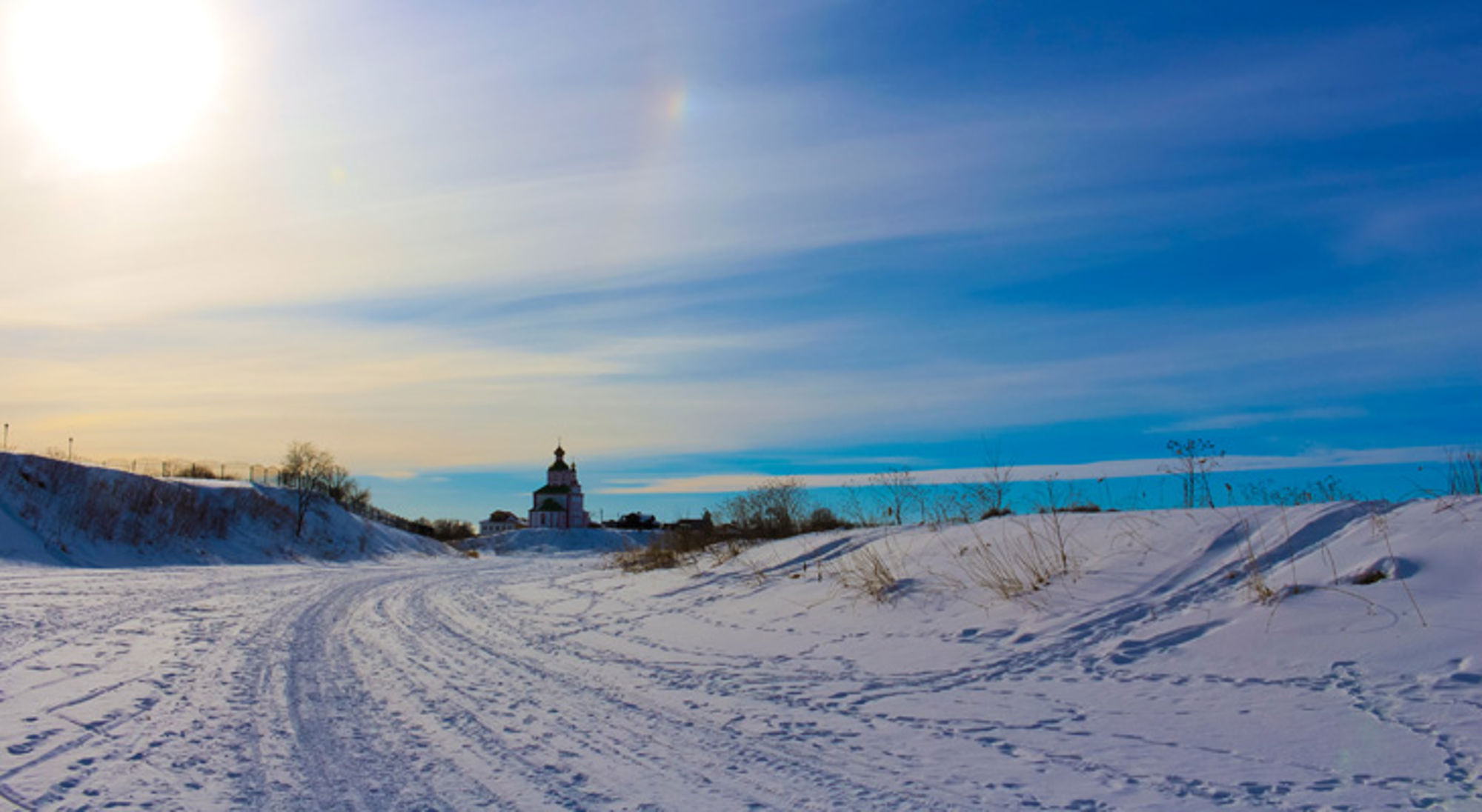 Besneeuwd landschap met kronkelend pad naar een kerk in de verte onder een blauwe winterlucht.