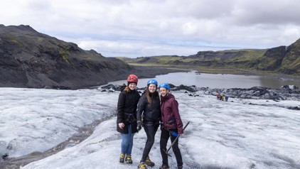 Drie reizigers met helm en stijgijzers tijdens een gletsjerwandeling in IJsland, met bergen en een meer op de achtergrond.