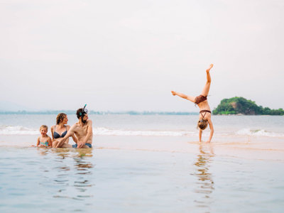 Gezin ontspant op het strand in Thailand, terwijl een kind een handstand doet bij de branding.